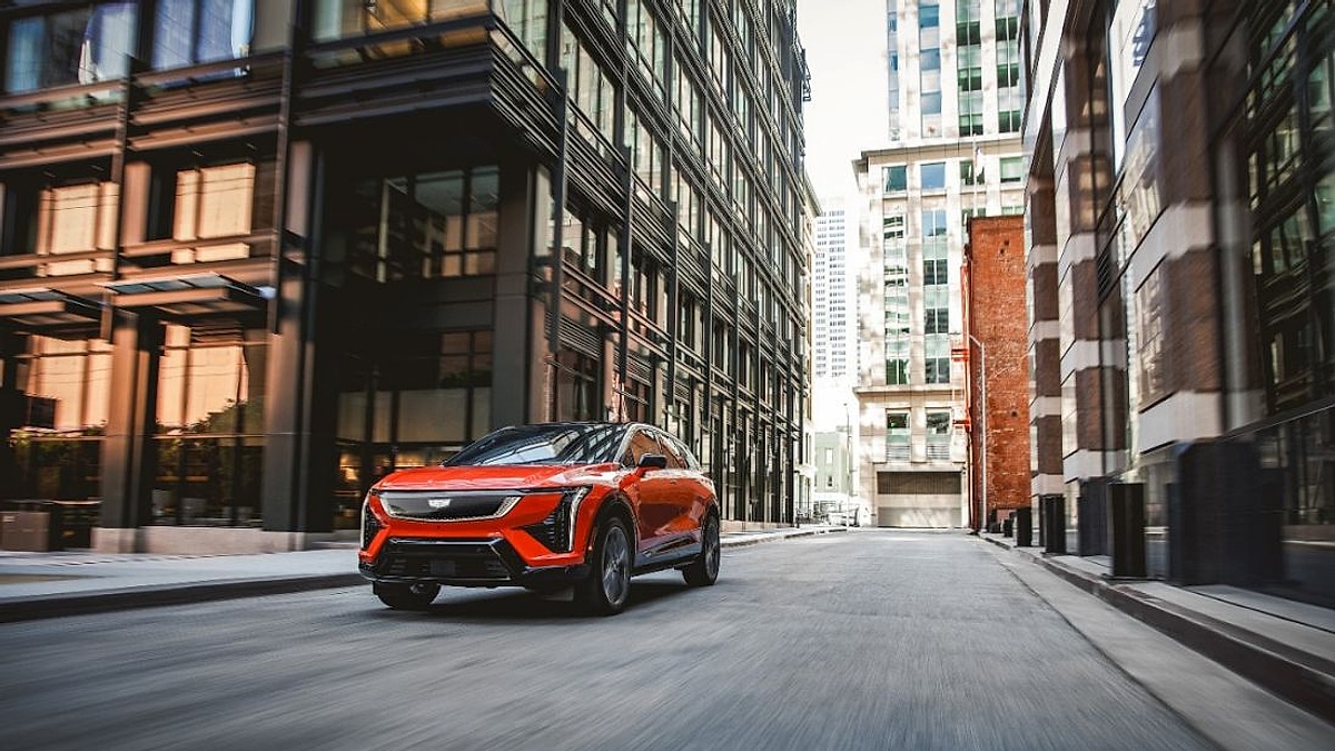 2025 Cadillac Optiq in Blaze Orange metallic accelerating through a narrow downtown city street flanked by glass skyscrapers with motion blur emphasizing the luxury electric crossovers commanding urban presence and performance